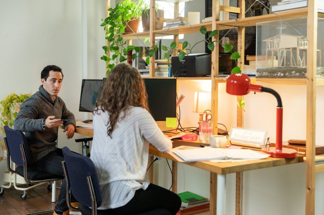 Man and woman talking at a shared wooden desk with monitors in a small design studio